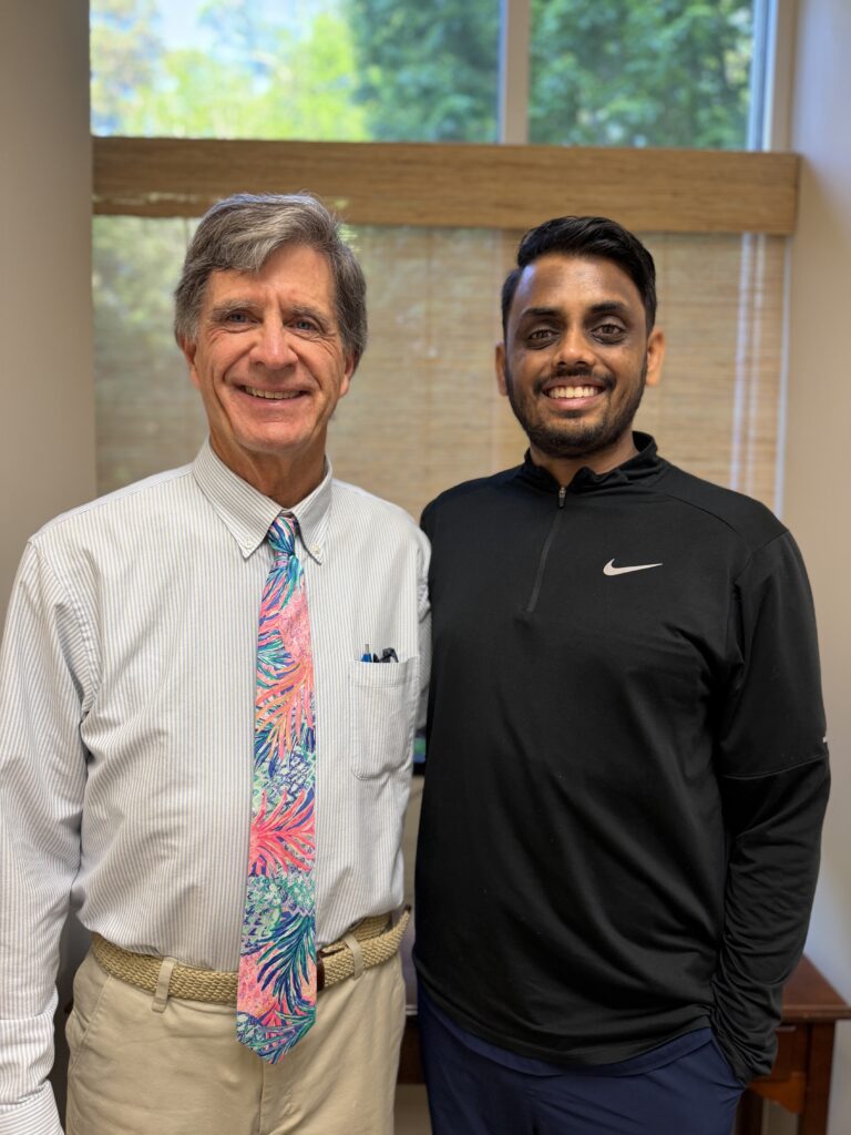 Two men standing indoors, smiling for the photo; left is an older man in a striped shirt and colorful tie, right is a younger man in a black athletic top.