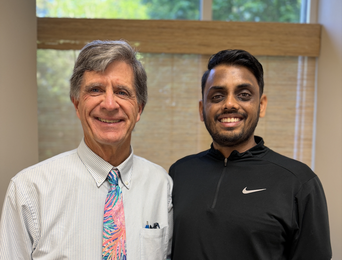 Two men stand side by side indoors, smiling at the camera: an older man in a striped dress shirt with a colorful tropical tie and pens in his pocket, and a younger man in a black Nike pullover.